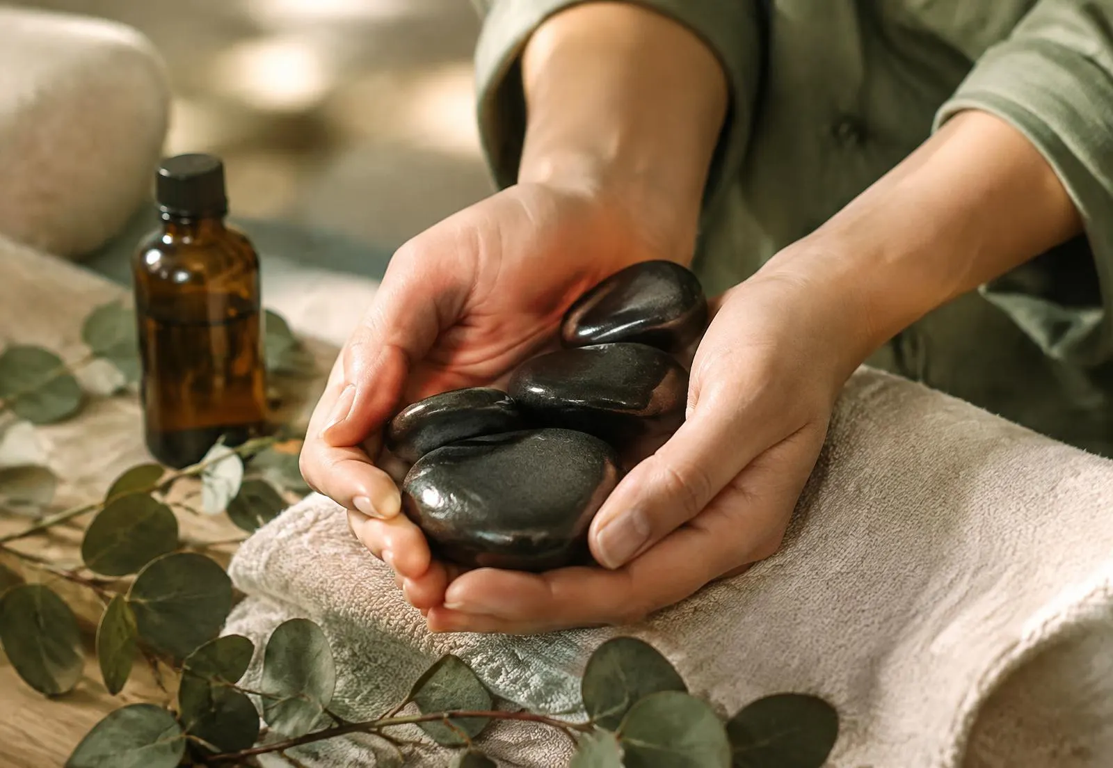Manos de terapeuta sosteniendo piedras volcánicas calientes junto a aceite esencial y eucalipto